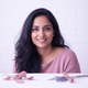 Portrait of a smiling South Asian woman in a dusty rose top against a bright white and lavender background.